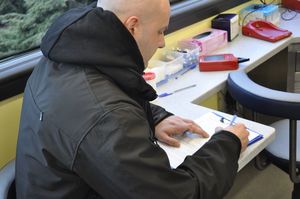 Na zdjęciu mężczyzna wypisuje formularz zgłoszeniowy honorowego dawcy krwi w ambulansie stojącym przed Komendą Wojewódzką Policji w Katowicach.