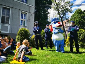 Policjanci rozmawiają z dziećmi. Obok nich stoi maskotka śląskiej policji. Dzieci siedzą na trawie.