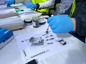 Na zdjęciu zbliżenie na odbitą na kartce dłoń. Technik kryminalistyki pokazuje w jaki sposób zabezpiecza się ślady daktyloskopijne.