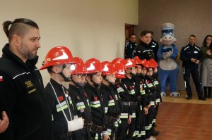 Młodzi strażacy stojący w szeregu, obok których stoi maskotka policji smoczyca Klara