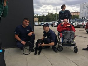 Policjant kuca przy psie, obok mężczyzna w mundurze i niepełnosprawny na wózku inwalidzkim.