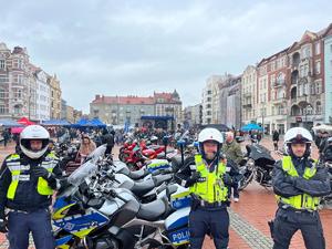 Na zdjęciu trzech policjantów, za nimi motocykle służbowe i zgromadzone na rynku osoby.