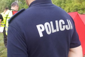 napis policja na koszulce , policjant odwrócony tyłem, w tle technik kryminalistyki i czerwony parawan