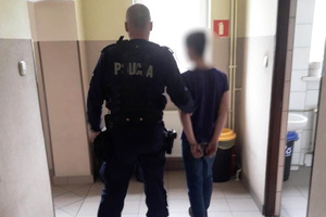 policjant prowadzi zatrzymanego, który ma kajdanki na rękach trzymanych z tyłu, zdjęcie w pomieszczeniu, na korytarzu