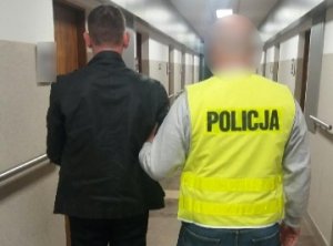 policjant w kamizelce odblaskowej z napisem POLIOCJA stoi tyłem na korytarzu trzymając pod rękę mężczyznę podejrzanego o rozboje