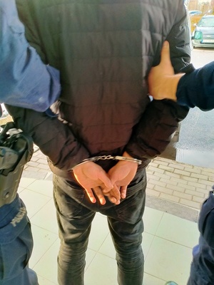 Zdjęcie zatrzymanego, który ma założone kajdanki oraz w części widocznych dwóch policjantów, którzy go prowadzą.