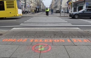 Na zdjęciu widać napis odłóż telefon i żyj naniesiony na przejście dla pieszych w tel widac jadące pojazdy oraz policjantów stojących przy przejściu