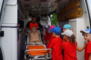 zdjęcie przedstawia karetkę pogotowia a w jej wnętrzu ratownika medycznego wraz z grupką dzieci i fantomem na noszach