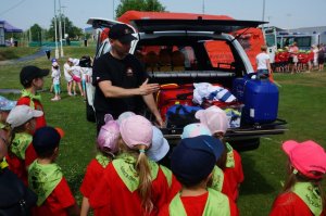 zdjęcie przedstawia strażaka pokazującego wyposażenie osobowego pojazdu strażackiego oraz przyglądającą się grupę przedszkolaków