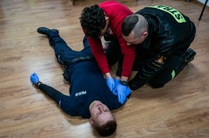 Zdjęcie - policjant w umundurowaniu leży na podłodze, przy nim klęczy strażak i mężczyzna (policjant) po cywilu. Strażak uciska miejsce symulowanej rany.
