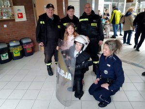 policjanta kuca przy dziewczynce, która założyła sprzęt którego policjanci używają na zabezpieczeniach. Obok jeszcze jedna dziewczynka i z tyłu 3 uśmiechniętych strażaków.
