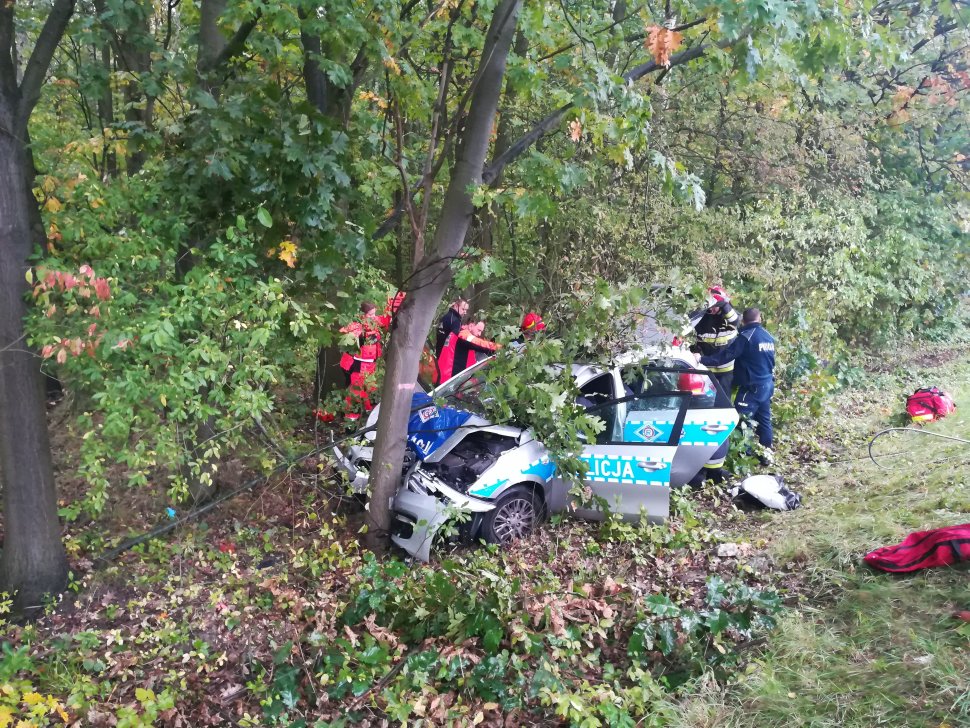 na zdjęciu widać kompletnie zniszczony radiowóz policyjny wbity w przydrożne drzewo. Obok radiowozu jest policjant, strażak oraz ratownicy medyczni