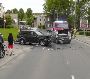 Zdjęcie przedstawia dwa rozbite samochody osobowe. W tle widać wóz strażacki, zaparkowane samochody.