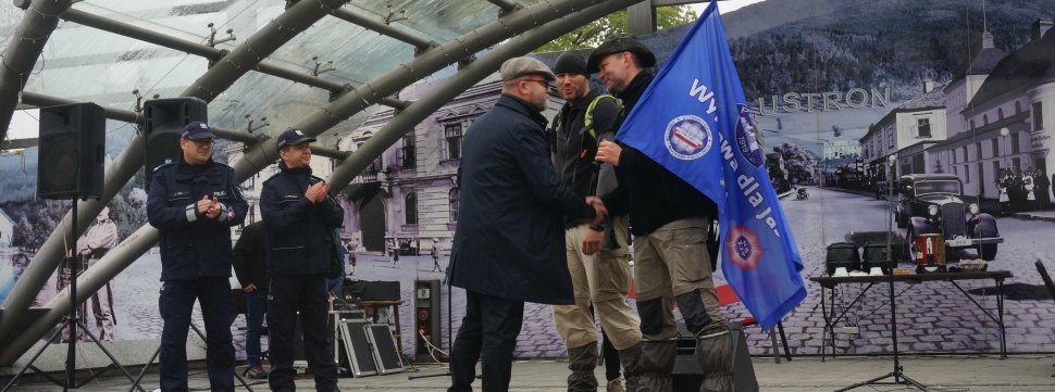 Obrazek przedstawia stojących na scenie na rynku w Ustronie po lewej stronie dwóch umundurowanych policjantów, któłrzy biją brawo, a z prawej strony burmistrza miasta Ustroń, który składa gratulacje dwóm nieumundorowanym policjantom z Tychów 