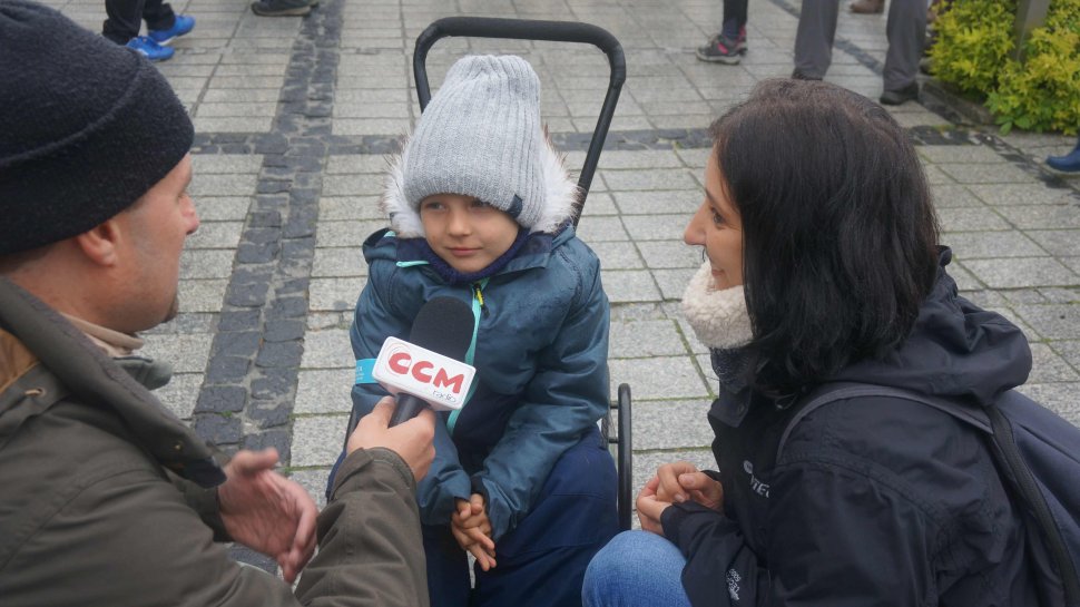 Obrazek przedstawia chłopca na wuzku inwalidzkim po przwej stronie jego mamę, a po lewej dziennikarza z radia CCM trzymającego przed chłopcem  mikrofon 
