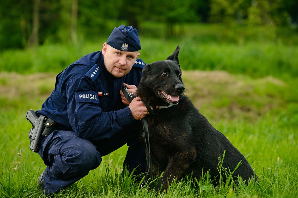 Na zdjęciu widoczny umundurowany policjant, który kuca i trzyma na smyczy psa służbowego. 