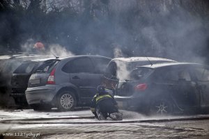 Obrazek przedstawia klęczącego strażaka, który dogasza zadymione pojazdy na parkingu