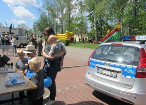 Obrazek przedstawia stojąca przy stoisku profilaktycznym policjantów kobietę z dziećmi, w tle widać policyjny radiowóz oraz zjeżdżalnię dla dzieci.