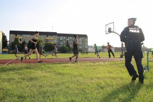 Na zdjęciu grupa osób uprawia sport na świeżym powietrzu, biegną jeden za drugim, na pierwszym planie widoczny także dzielnicowy w mundurze