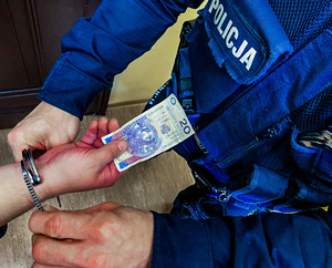 Na zdjęciu osoba  podaje banknot i ma zakładane kajdanki na ręce.