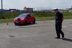 Zdjęcie czerwonego samochodu osobowego na placu manewrowym. Z boku stoi policjant.