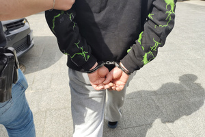 Na zdjęciu widoczny prowadzony mężczyzna, który na ręce trzymane z tyłu ma założone kajdanki.