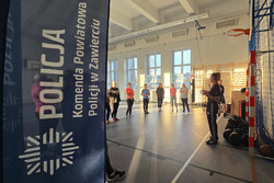Na zdjęciu widoczna z lewej strony flaga reklamowa z napisem Policja Komenda Powiatowa Policji w Zawierciu oraz kobiety słuchające instruktorki.