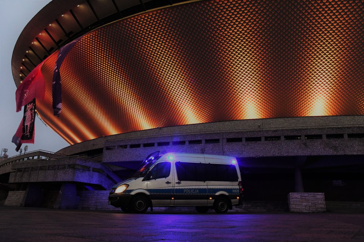 Oznakowany radiowóz policji z włączonymi światłami pojazdu uprzywilejowanego z budynkiem Katowickiego Spodka w tle.