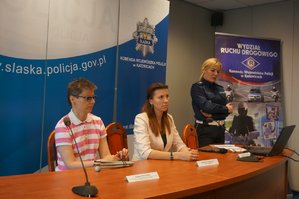 Na zdjęciu widać siedzących przy stole prelegentów oraz policjantka w czasie spotkania z mediami w siedzibie Komendy Wojewódzkiej Policji w Katowicach.
