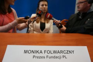 Zdjęcie przedstawia na pierwszym planie tabliczkę z napisem: Monika Folwarczny - Prezes Fundacji PL, a w tle siedzą dziennikarze wraz z Moniką Folwarczny.