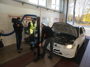Na zdjęciu samochód osobowy stojący na stacji diagnostycznej w czasie kontroli ustawienia świateł. Z lewej strony pojazdu stoi umundurowany policjant a za nim operator z kamerą.