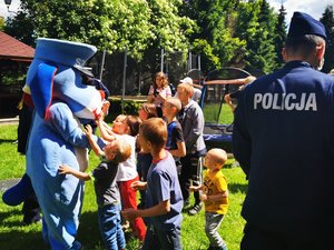 Dzieci bawią się z policyjną maskotką. Na pierwszym planie napis policja umieszczony na mundurze policjanta stojącego tyłem.