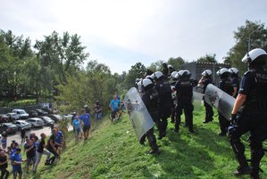 Grupa osób dobiega do policjantów w kaskach i kamizelkach, mundurowi trzymają tarcze.