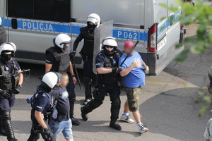 Umundurowani policjanci w kaskach i kamizelkach prowadzą dwóch mężczyzn.