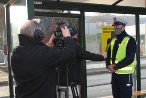 Zdjęcie kolorowe. Teren przystanku autobusowego. Umundurowany policjant udziela wywiadu dziennikarzowi przed kamerą