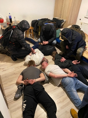 Zatrzymani przez policjantów mężczyźni leżą na podłodze w mieszkaniu, mają na dłoniach kajdanki. Obok nich nieumundurowani policjanci.