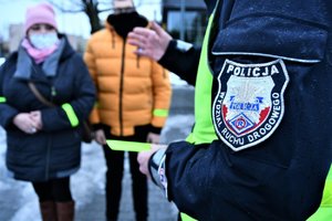 Policjant Wydziału Ruchu Drogowego Komendy Wojewódzkiej Policji w Katowicach trzyma w dłoniach odblask i tłumaczy pieszym jak należy dbać o bezpieczeństwo.