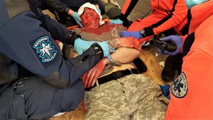Osoba odgrywająca poszkodowanego leży na podłodze. Policyjny ratownik oraz medycy udzielają pierwszej pomocy.