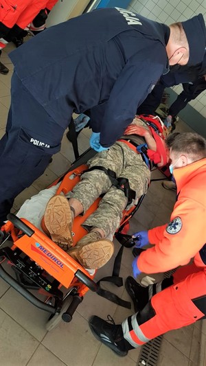 Osoba odgrywająca poszkodowanego leży na noszach. Policyjny ratownik oraz medycy udzielają pierwszej pomocy.