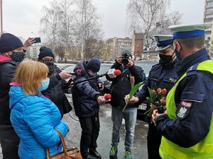 Na zdjęciu policjant oraz członek kabaretu przebrany w policyjny mundur, trzymający tulipany. Jednego z nich wręcza stojącej naprzeciwko kobiecie. Obok stoją przedstawiciele mediów, trzymający mikrofony. Jeden z mężczyzn robi zdjęcia.