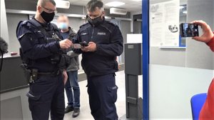 Na zdjęciu umundurowani policjanci podczas legitymowania, obok widoczna ręka mężczyzny, który telefonem nagrywa przebieg interwencji.