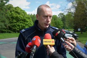 Zdjęcie kolorowe. Umundurowany policjant na terenie Parku Śląskiego wypowiada się do mikrofonów i kamer reporterów.