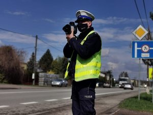 Umundurowany policjant na drodze, trzymający urządzenie służące do pomiaru prędkości z jaką poruszają się samochody.