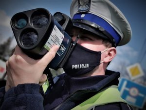 Zbliżenie na twarz policjanta w maseczce ochronnej, który przy użyciu laserowego miernika prędkości sprawdza prędkość, z jaką poruszają się kierowcy.