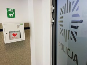 Skrzynka z napisem AED, w której znajduje się defibrylator. Na szklanych drzwiach napis Policja. Zdjęcie wykonane w budynku komendy Policji.