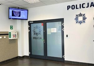 Skrzynka z napisem AED, w której znajduje się defibrylator. Na szklanych drzwiach i ścianie widnieje napis Policja, na drugiej ścianie zamontowany ekran informacyjny, który wyświetla naszywkę na policyjnym mundurze. Zdjęcie wykonane w budynku komendy Policji.