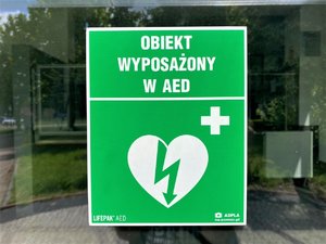 Zielona tabliczka na szklanych drzwiach z napisem obiekt wyposażony w AED. Pod napisem białe serce z błyskawicą na środku oraz krzyżyk.