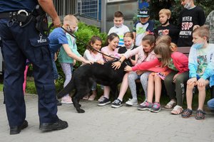 Zdjęcie kolorowe. Widoczny policjant przewodnik psa służbowego oraz jego pies służbowy, a także uczestnicy spotkania, głównie dzieci
