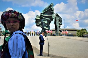 Dziewczynka ubrana w regionalny, kolorowy strój odwraca się w stronę fotografa. W tle Pomnik Powstańców Śląskich.