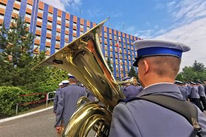 Zdjęcie kolorowe. Widoczny umundurowany muzyk policyjne orkiestry trzymający instrument dęty, w tle inni policjanci biorący udział w uroczystości oraz budynek komendy wojewódzkiej policji